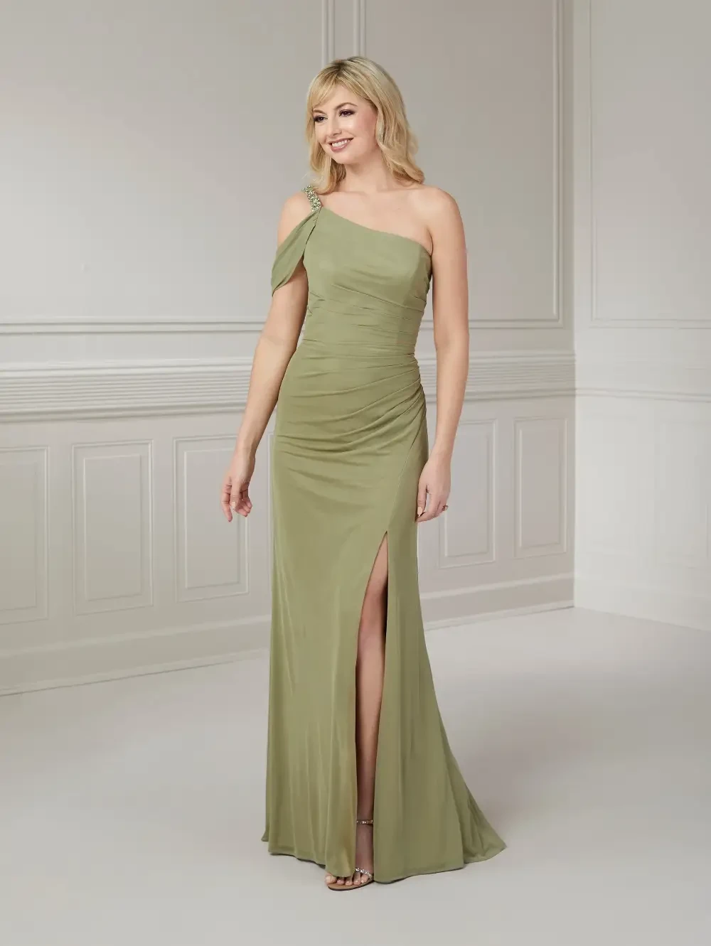 A woman wearing a one-shoulder olive green gown with ruching and a thigh-high slit, standing in a well-lit room with white walls.