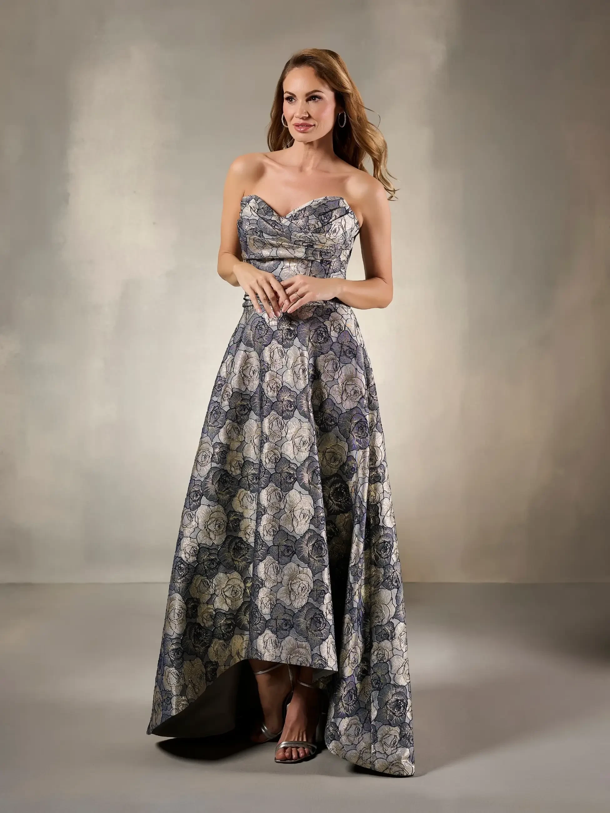 A woman wearing a strapless, patterned evening gown with a high-low hem, standing against a neutral background.