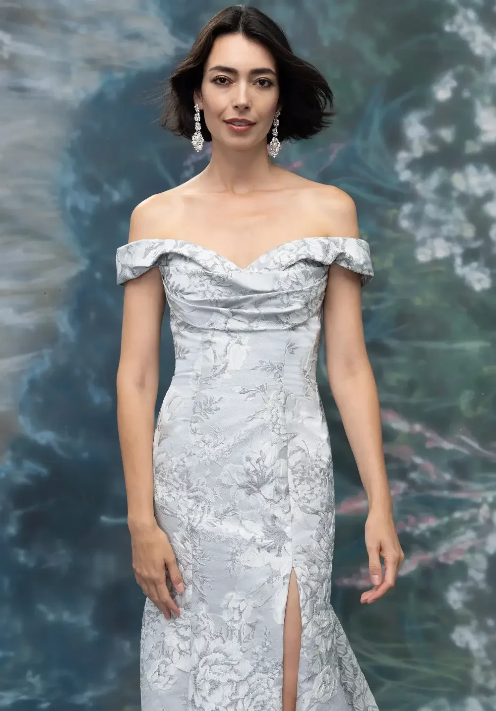 A woman wearing an off-the-shoulder, silver floral gown with a thigh-high slit, standing against a blurred background of soft colors.