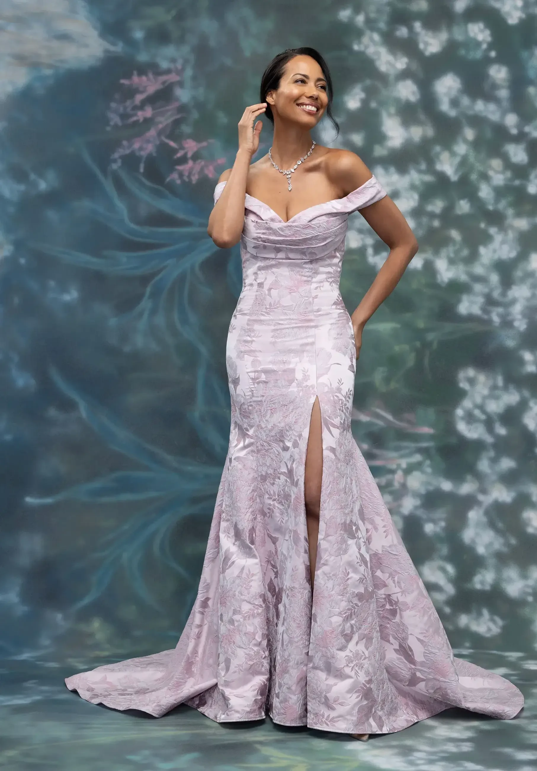 A woman is wearing an elegant lavender evening gown with off-the-shoulder sleeves. The dress features a thigh-high slit and intricate floral patterns, and she is accessorized with a necklace. She stands against a soft, blurred background of flowers.