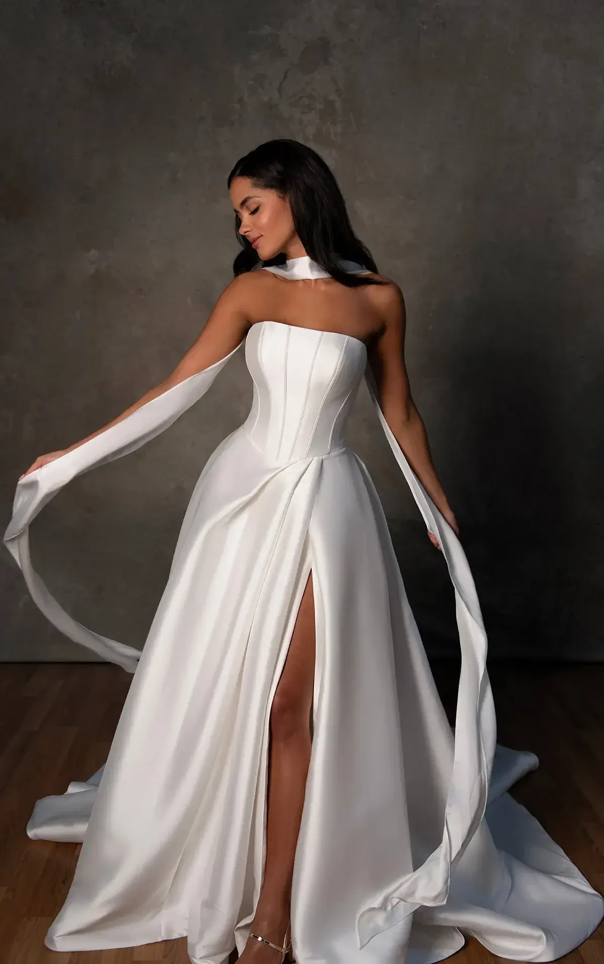A woman in an elegant, strapless white gown with a high slit stands on a wooden floor. The dress features long, flowing ribbons, and she exudes grace and confidence against a muted backdrop.