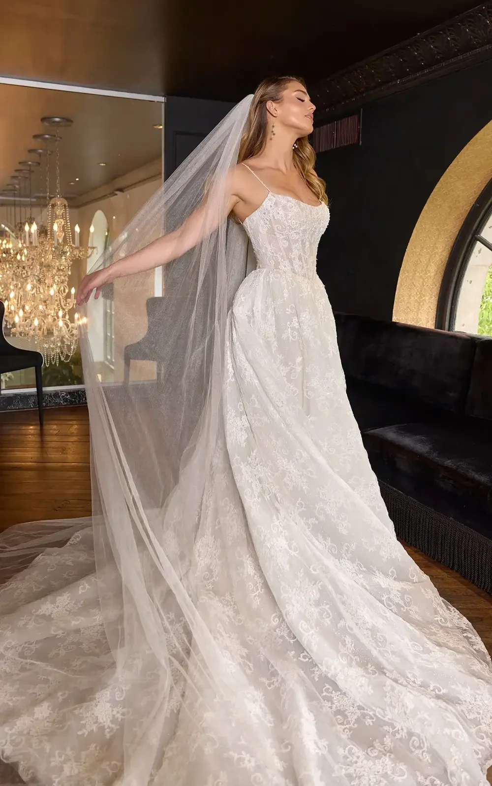 A bride in a stunning white wedding dress with intricate lace details, gracefully wearing a long veil, striking a pose in an elegant indoor setting.