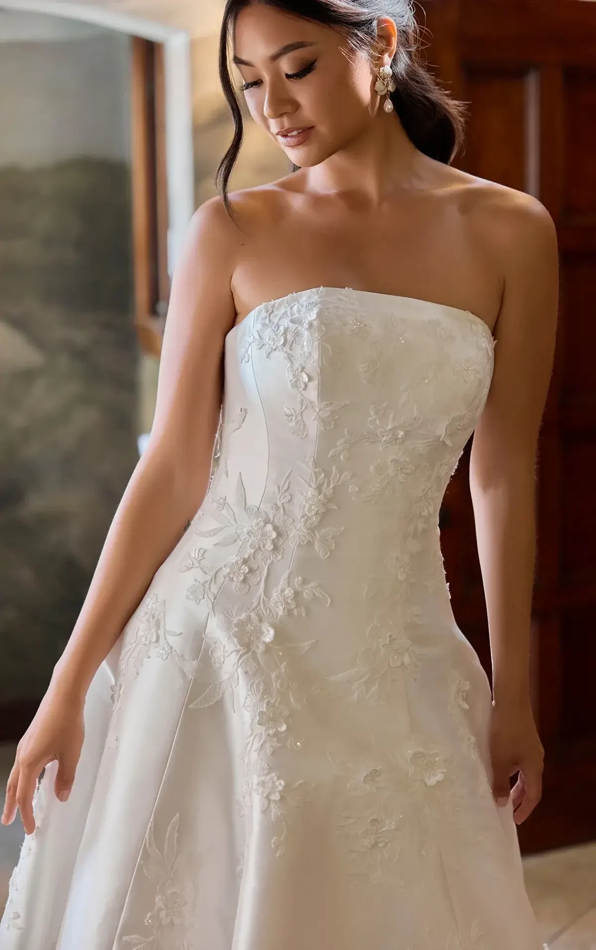 A woman in a strapless, white wedding dress with floral embroidery, posing gracefully.