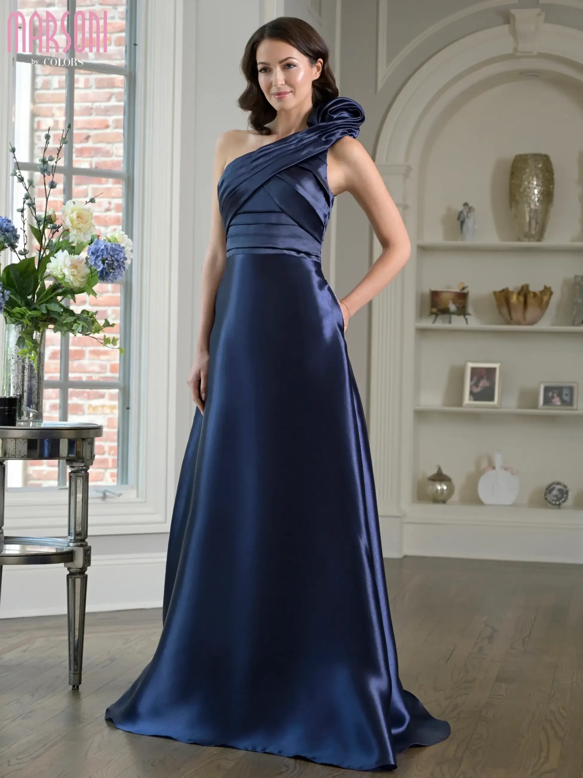 A woman wearing a navy blue, one-shoulder formal gown, standing elegantly in a well-decorated room with flowers and tasteful decor in the background.