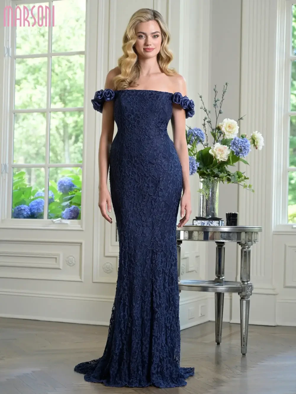 A woman in a fitted off-the-shoulder navy blue gown with floral accents, standing elegantly in a bright room adorned with flowers and decor.
