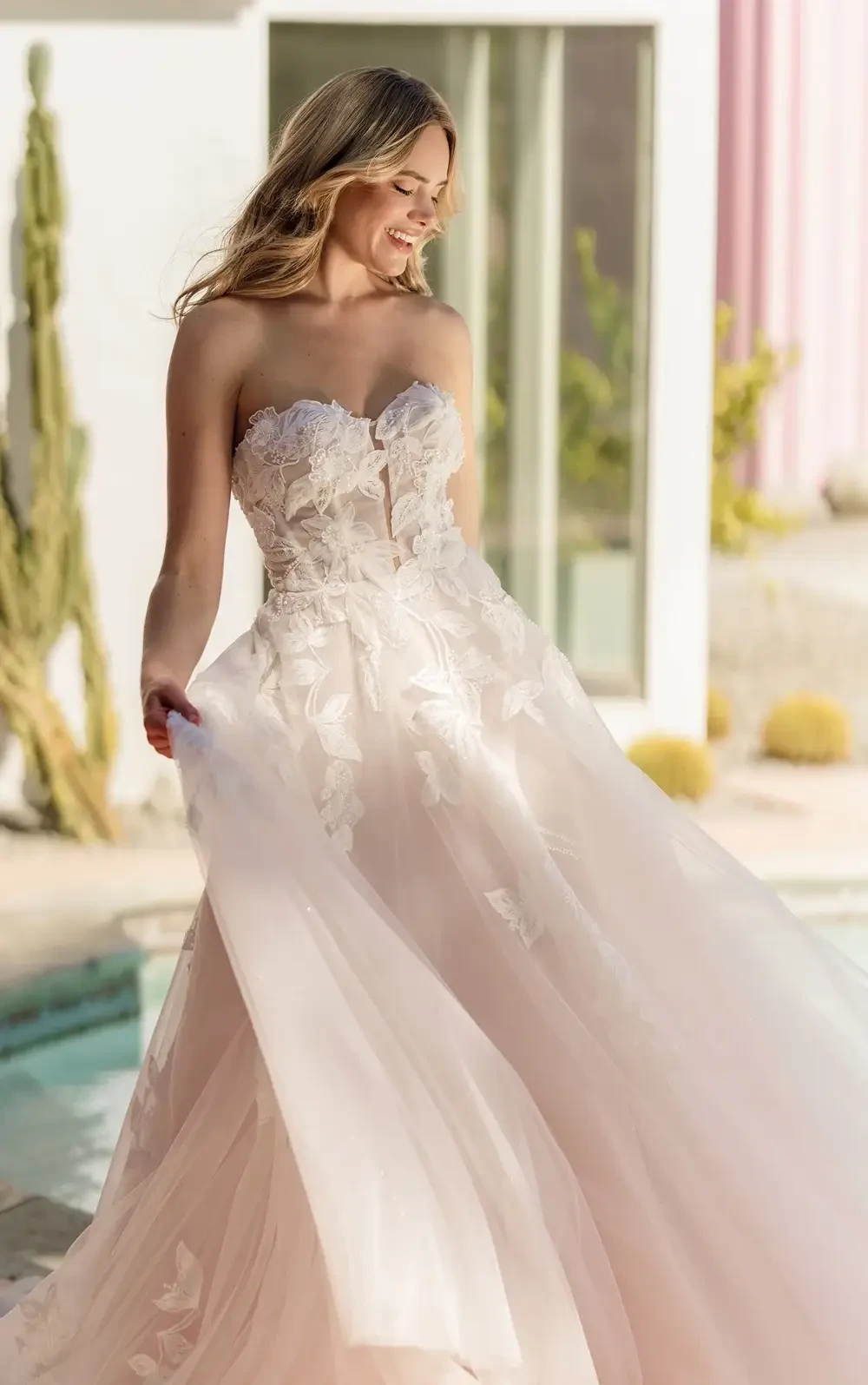 A woman in a strapless, floral-embellished gown twirls gracefully outdoors, with a soft, flowing skirt and a joyful expression.
