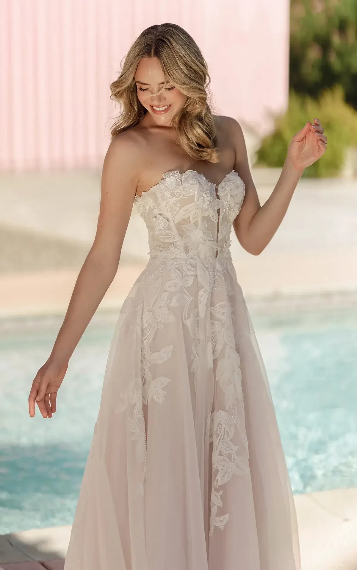 A woman in a strapless, flowy gown with floral patterns, standing by a pool and smiling.