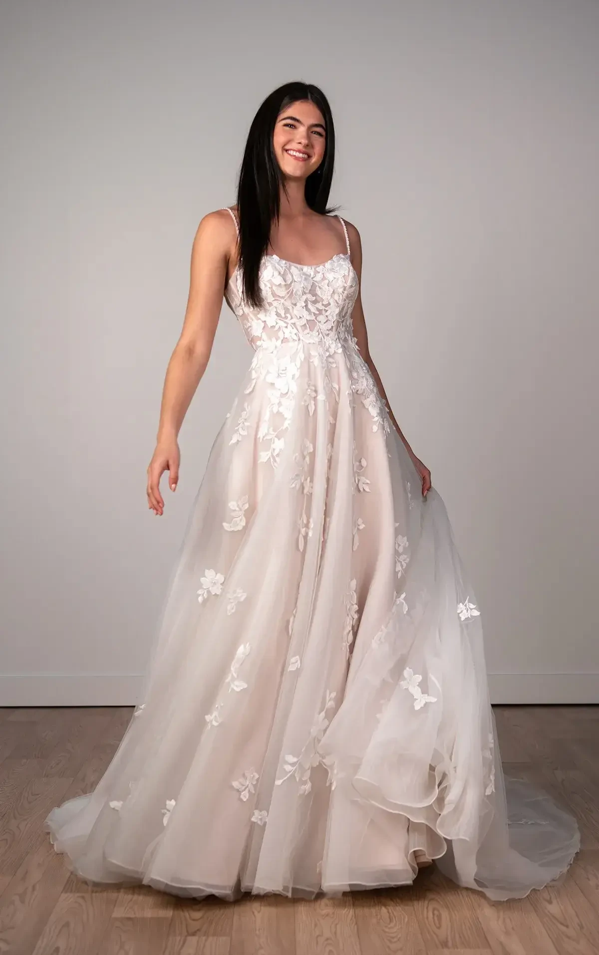 A smiling woman wearing a flowing, elegant wedding dress with floral designs stands in a well-lit room.
