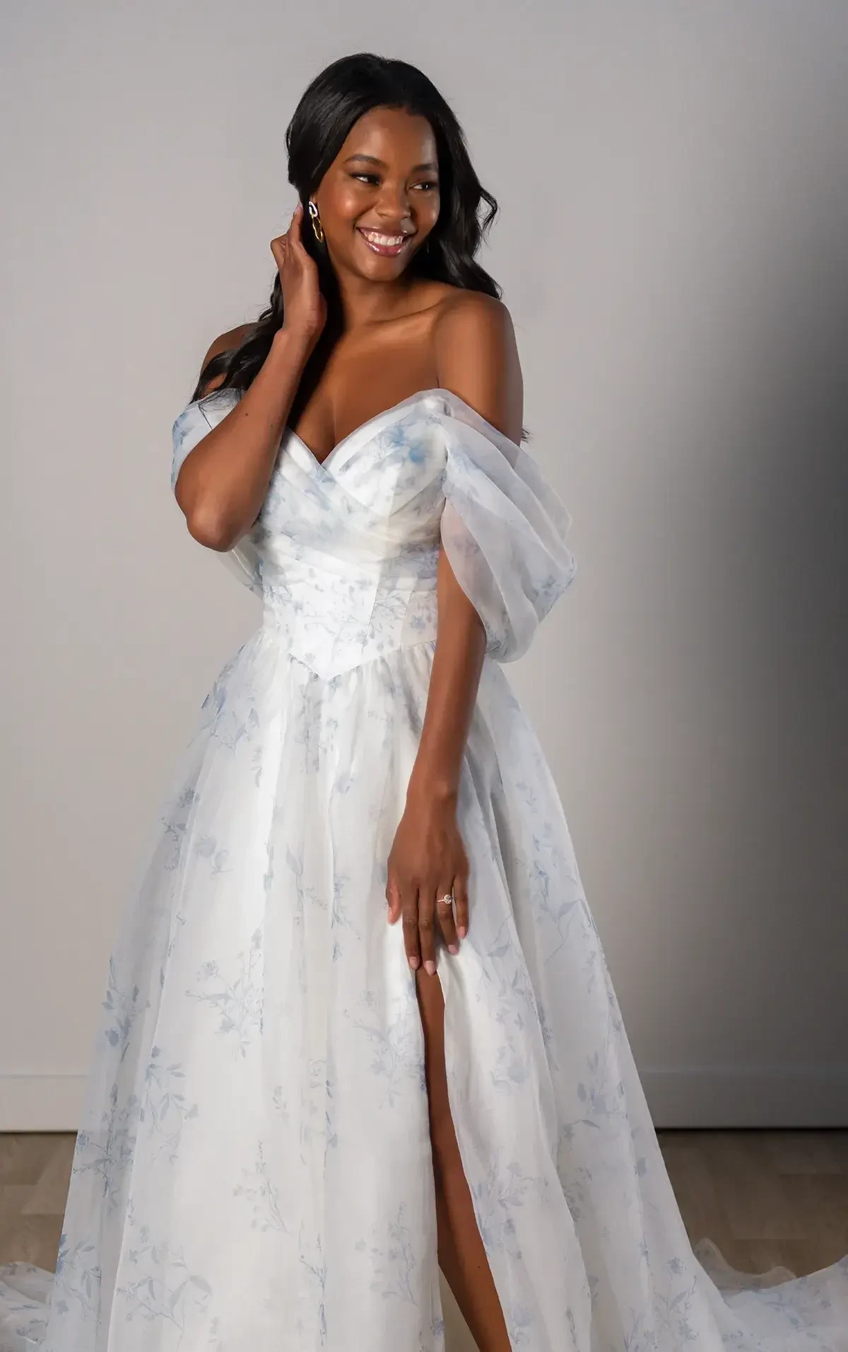 A woman wearing a beautiful white gown with off-the-shoulder sleeves, smiling and posing in a well-lit setting. The gown has a thigh-high slit and features delicate detailing.